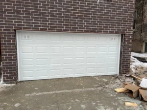 A wide, white double-car garage door with a raised panel design and corner ventilation inserts installed in a dark brick facade.